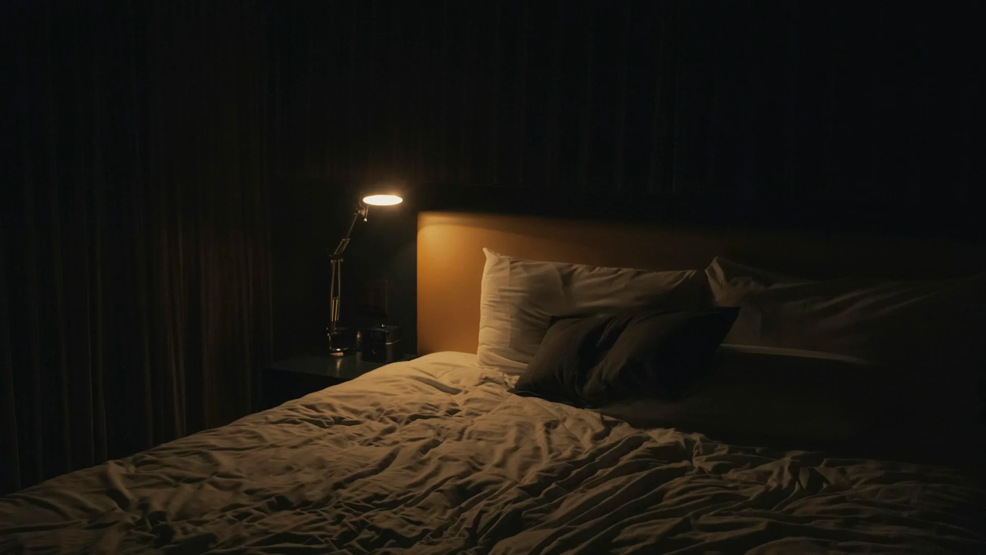 Empty made bed glowing under a warm bedside lamp in a dark, cozy bedroom at night