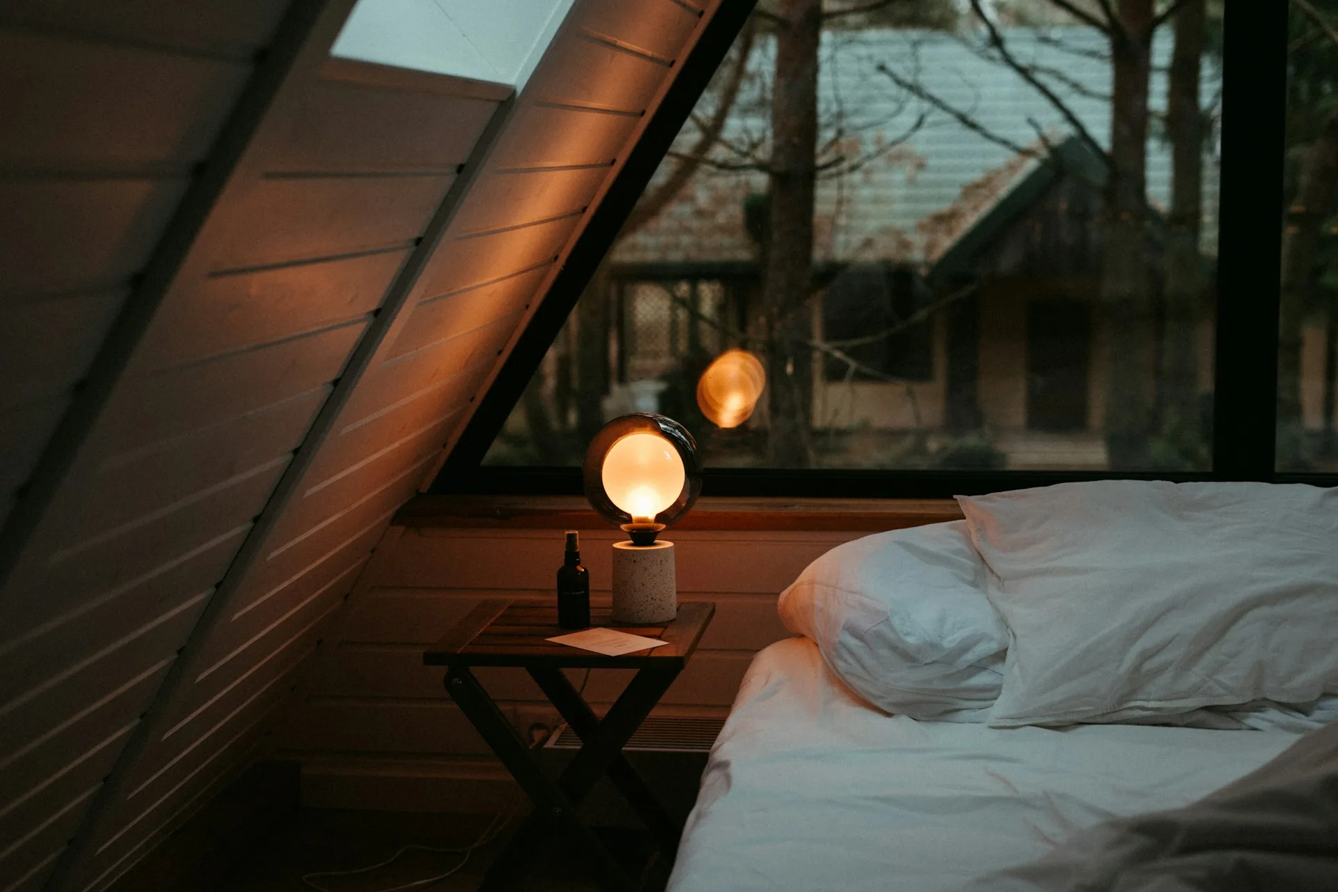 A cozy bed beside a warm bedside lamp and a triangular cabin window at night