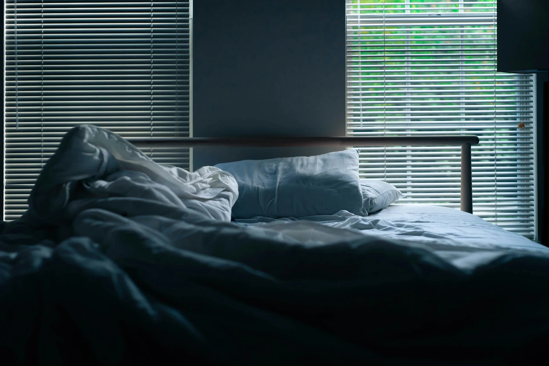 An empty, unmade bed lit by soft morning light through window blinds