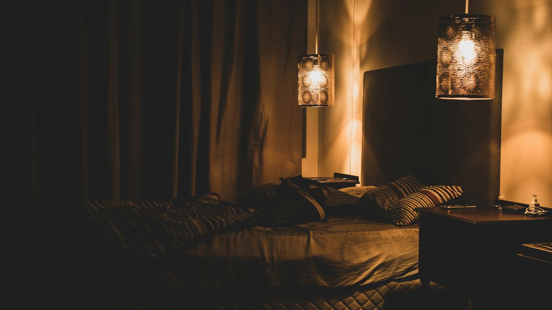 Two pendant lamps glowing over a dimly lit bed with rumpled bedding and striped pillows at night