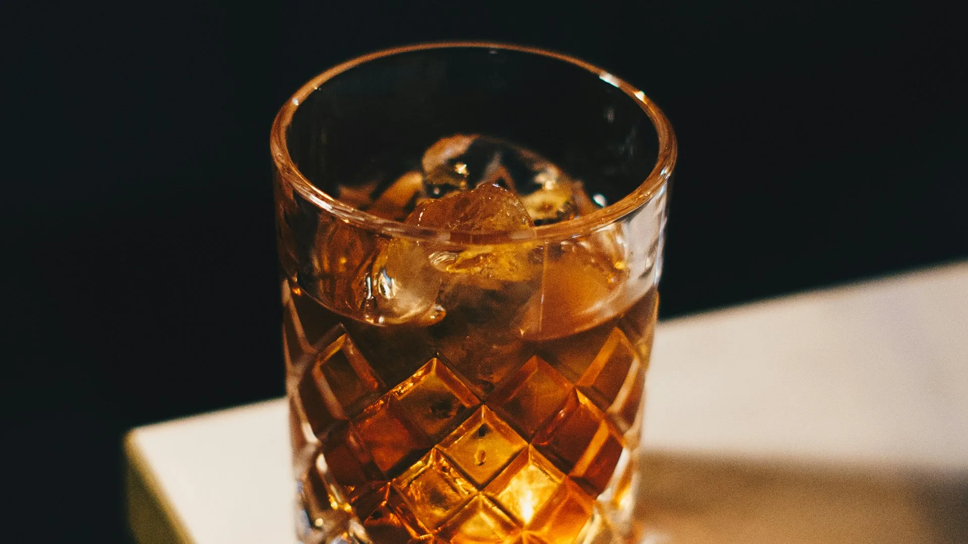 Cut-crystal whiskey glass with ice on a dark marble table, warm low-key lighting
