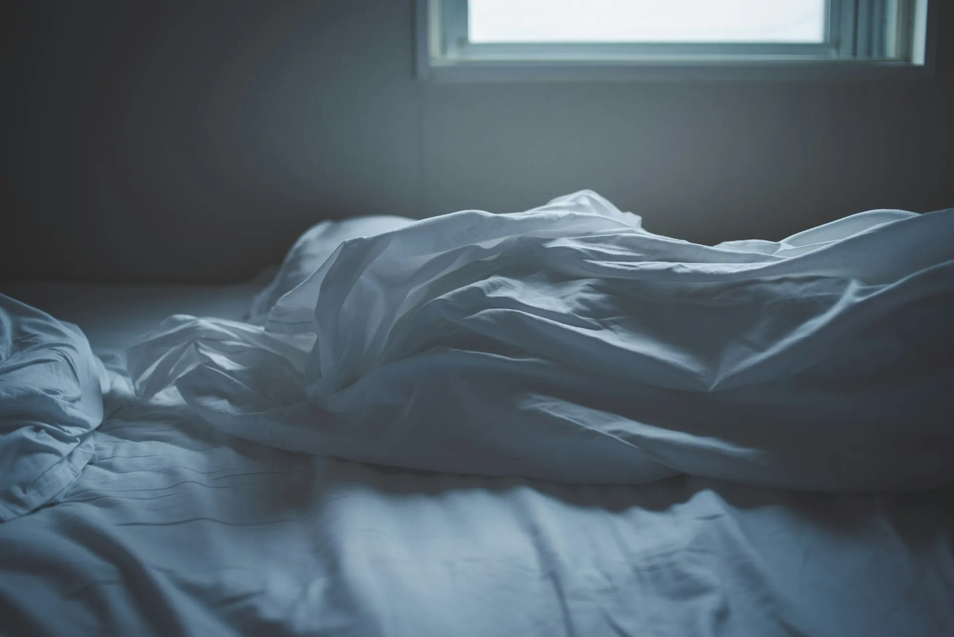 An empty, rumpled bed lit by cool morning light through a small window
