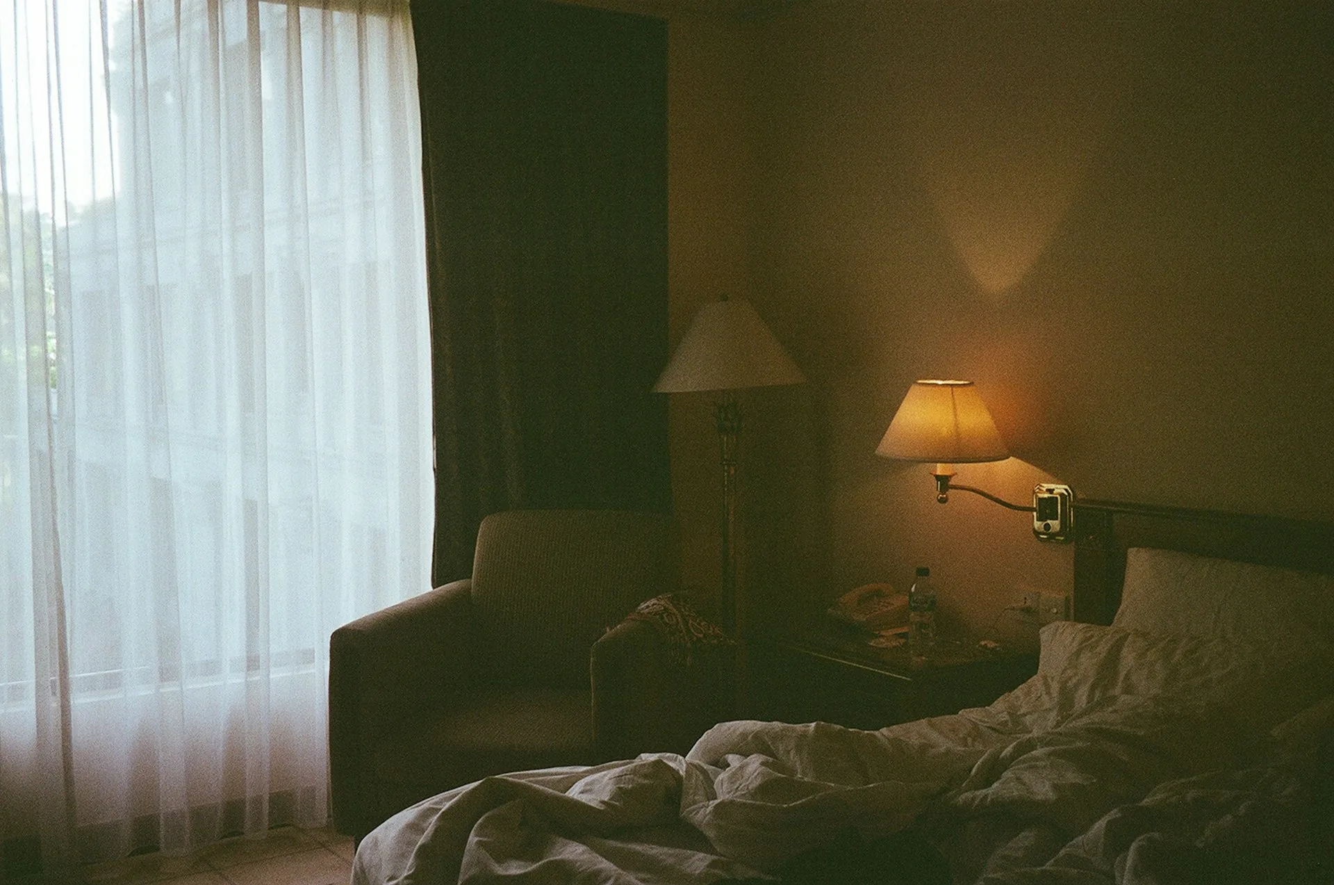 An empty, unmade hotel bed beside a warm lamp and a sheer curtained window
