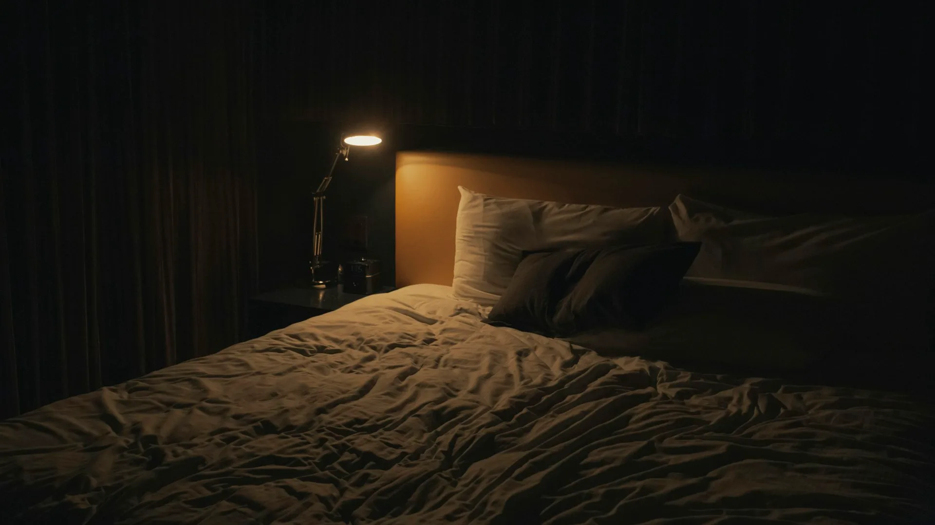 Warm amber bedside lamp glowing beside a dark, quiet bed at night