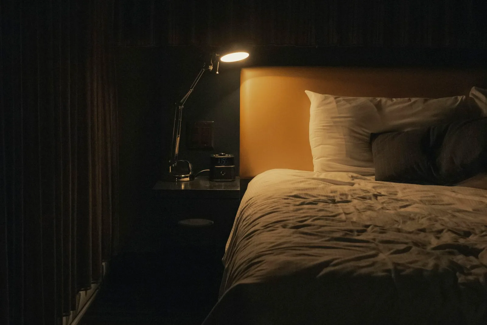 Warm study lamp glowing beside an unmade bed in a dark, quiet bedroom at night