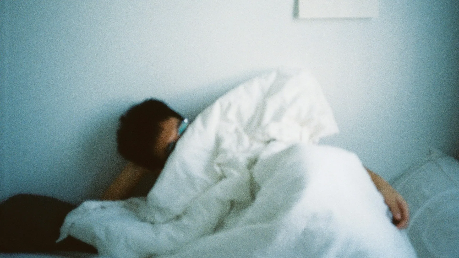 Person resting under a white blanket in a dim, still bedroom